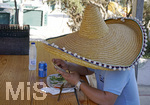 06.09.2017, Spanien, Insel Mallorca,  Parc natural de Mondrag, Cala Mondrago. Strand bei Mondrago. Ein Urlauber schaut auf sein Handy whrend des Essens in einer Strandbar, versteckt unter einem riesigen Sombrero.
