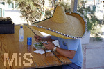 06.09.2017, Spanien, Insel Mallorca,  Parc natural de Mondrag, Cala Mondrago. Strand bei Mondrago. Ein Urlauber schaut auf sein Handy whrend des Essens in einer Strandbar, versteckt unter einem riesigen Sombrero.