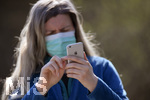 08.04.2020, Symbolbild, Corona-Krise, Frau schaut auf ihr Handy mit  Gesichtsmaske.