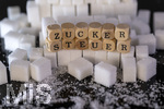 14.10.2026, Thema Zuckersteuer für Softgetränke. Studioaufnahme mit Würfelzucker und Schriftzug ZUCKERSTEUER.  Die Bundesregierung hat sich im Streit um die Zuckersteuer für Getränke geeinigt, Der Kompromiss sieht eine zweistufige Steuer und Ausnahmen für kleinere Hersteller vor. So soll ab dem 1. Juli 2027 ein Steuersatz von 26 Cent auf gesüßte Getränke mit einem Zuckergehalt ab 5 Gramm pro 100 Milliliter erhoben werden. Bei einem Zuckergehalt von 8 Gramm pro 100 Milliliter soll der Satz 32 Cent betragen.